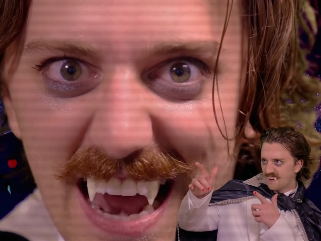 Brian David Gilbert, dressed as a vampire, bares his fangs at the camera. In the bottom right corner, a smaller version of him shoots finger guns.